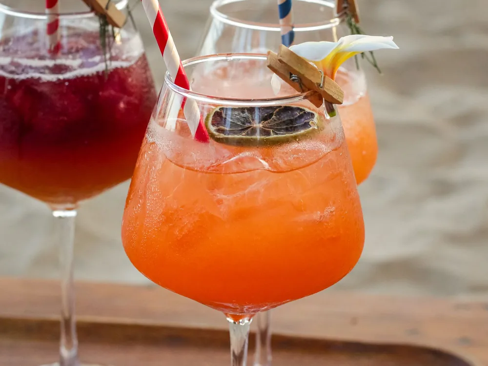 Two colourful cocktails with straws and fruit garnish in close-up, with a soft beach behind