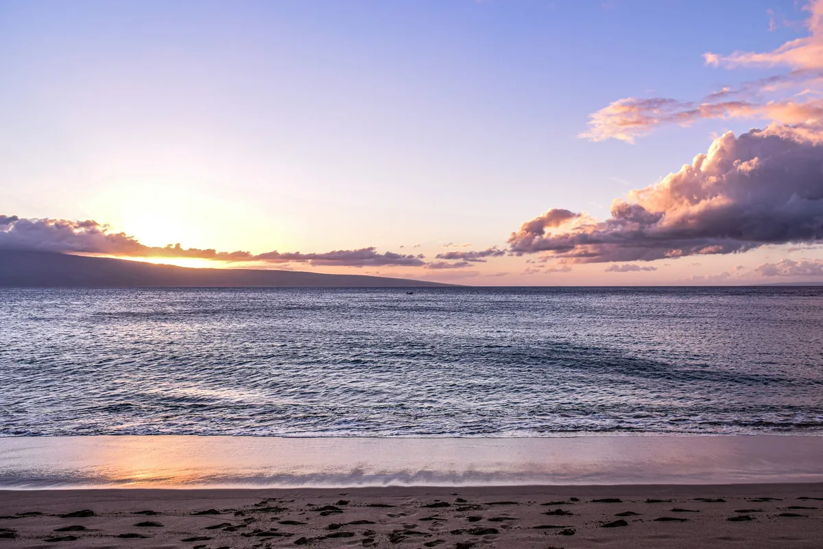 The sun low over a calm sea with the dark outline of West Maui and soft violet clouds above